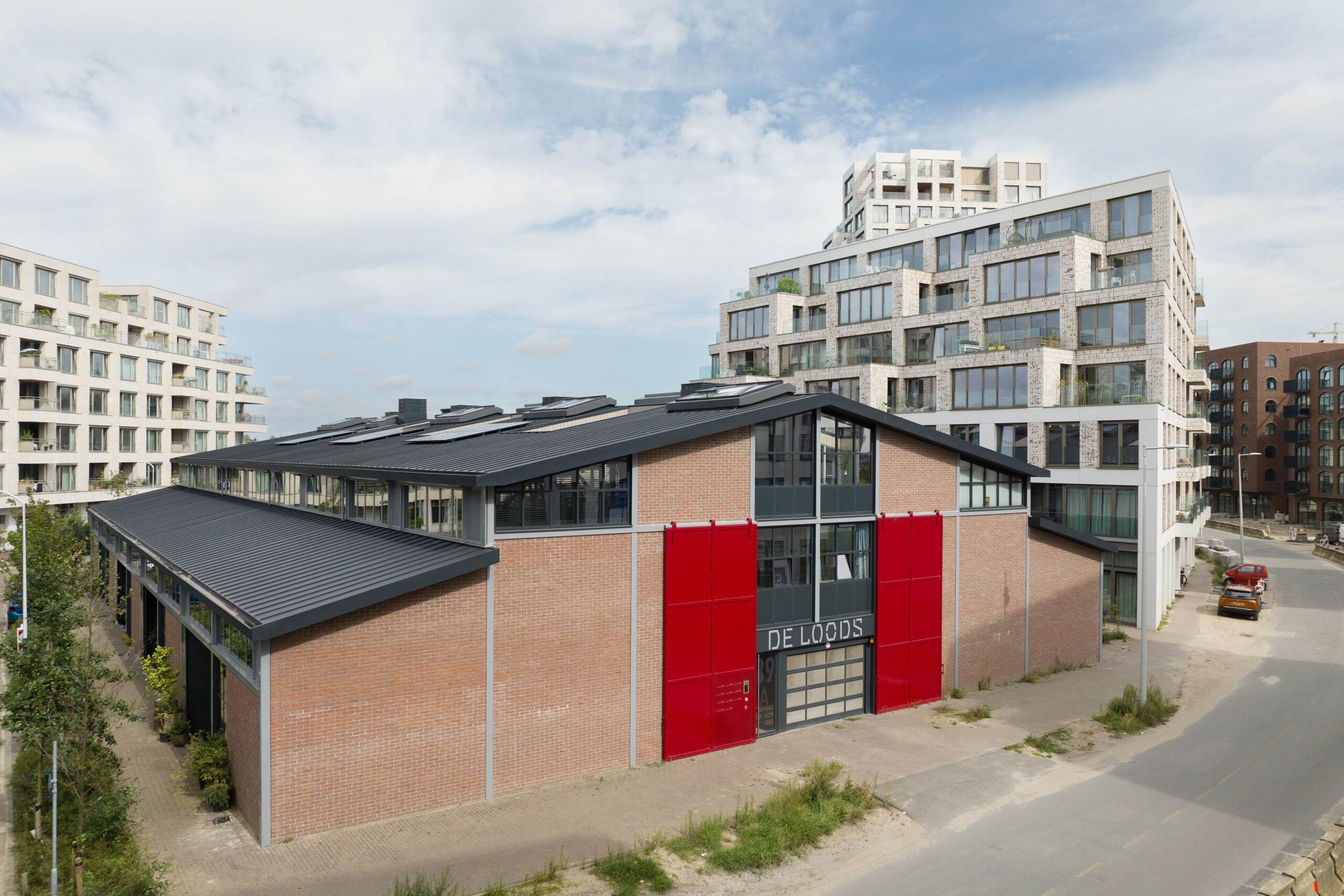 Gebouw "De Loods" met rode deuren en appartementencomplexen op de achtergrond.