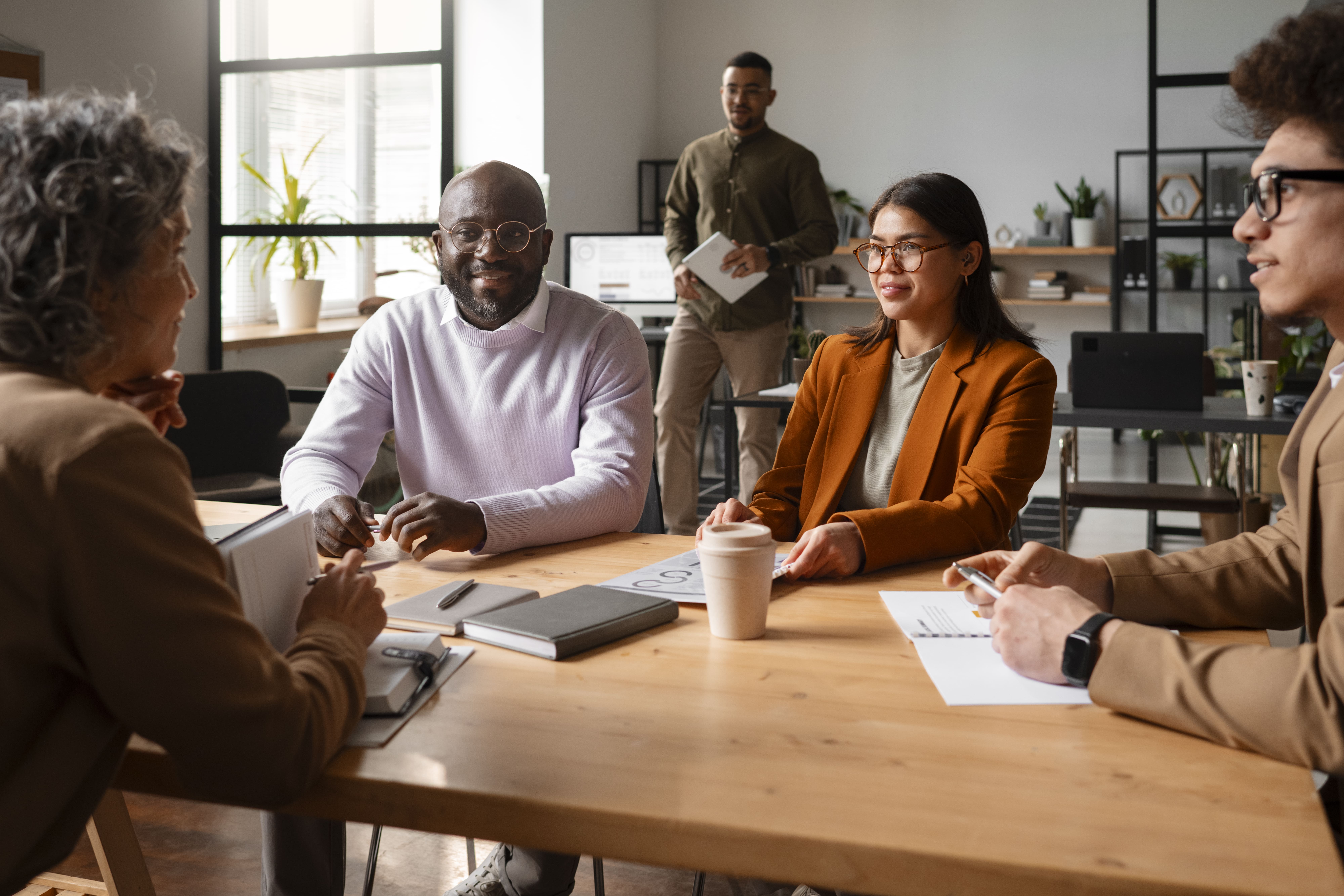 A imagem mostra um grupo diversificado de colegas em uma reunião de trabalho, sentados ao redor de uma mesa de madeira em um ambiente de escritório moderno e iluminado. A cena transmite colaboração e informalidade, com os integrantes usando roupas casuais e de negócios. A variedade de idades e etnias representadas enfatiza a diversidade no ambiente corporativo. A interação entre os membros do grupo, com expressões amigáveis, sugere um ambiente inclusivo e de diálogo aberto, ideal para discussões produtivas e trabalho em equipe.