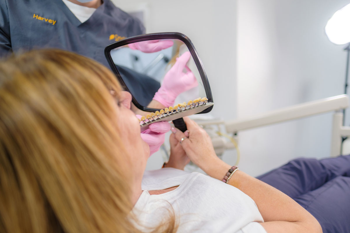 A female dental patient with blonde hair reclines in a dental chair, holding a hand mirror and a tooth shade guide with various small, tooth-colored tabs. She is comparing the different shades to her own teeth, while a dentist wearing blue scrubs with the name "Harvey" and pink gloves assists her from behind.