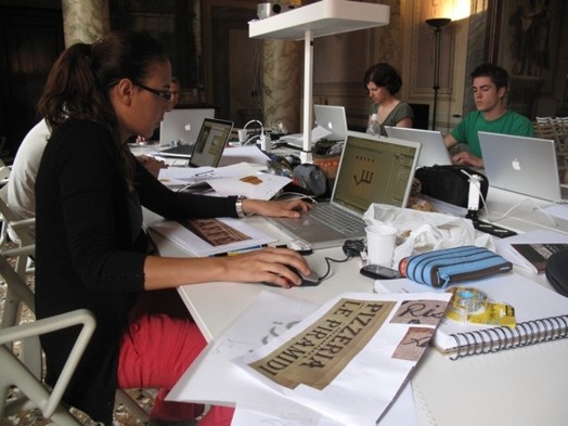 A photo of several students working on laptops at a table while in the middle there is a projector.