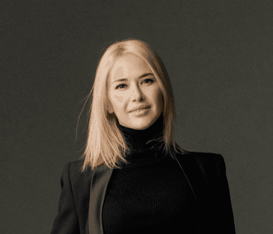 A person with shoulder-length blonde hair and a confident expression is wearing a black turtleneck and blazer, standing against a neutral background.