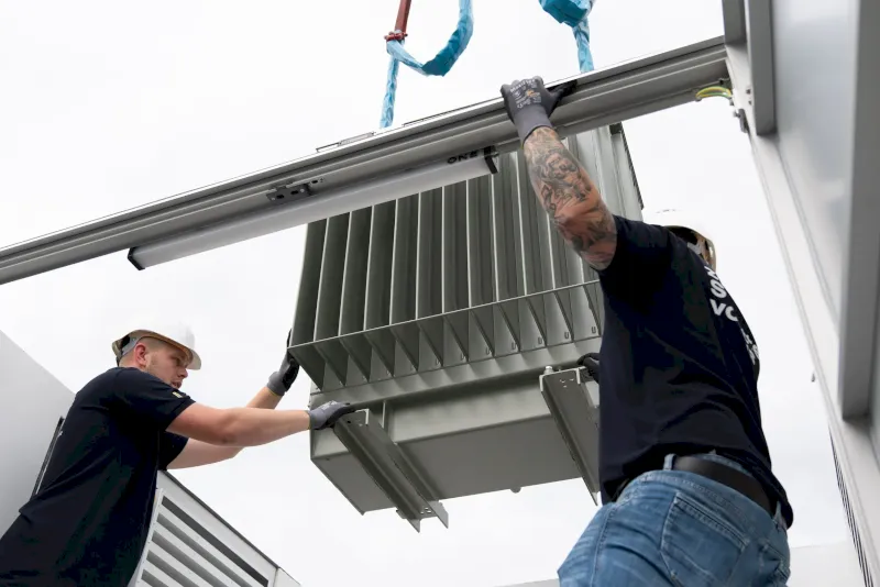 Zwei Arbeiter heben mit einem Kran einen großen Transformator.