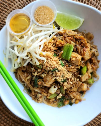 A white bowl of Phad Thai noodles with tofu, green onions, and crushed peanuts, served with fresh bean sprouts, lime, and traditional condiment cups.