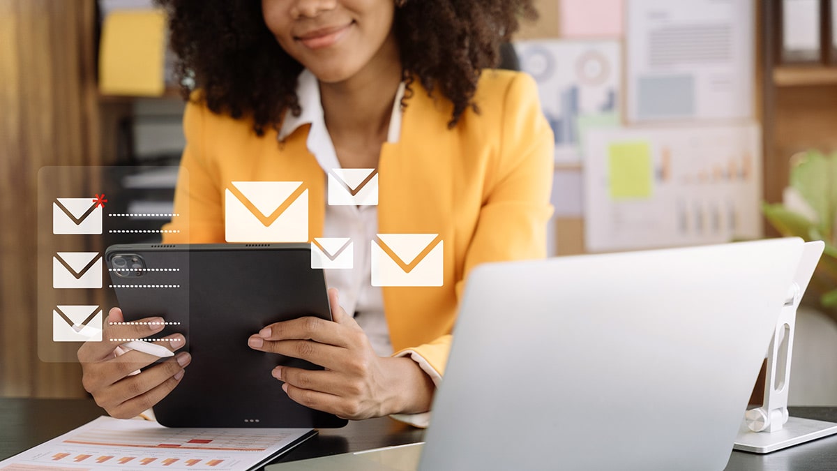 A woman in a bright yellow blazer is seated at a desk, holding a tablet while surrounded by floating email icons, signifying digital communication and connectivity in a modern office setting.