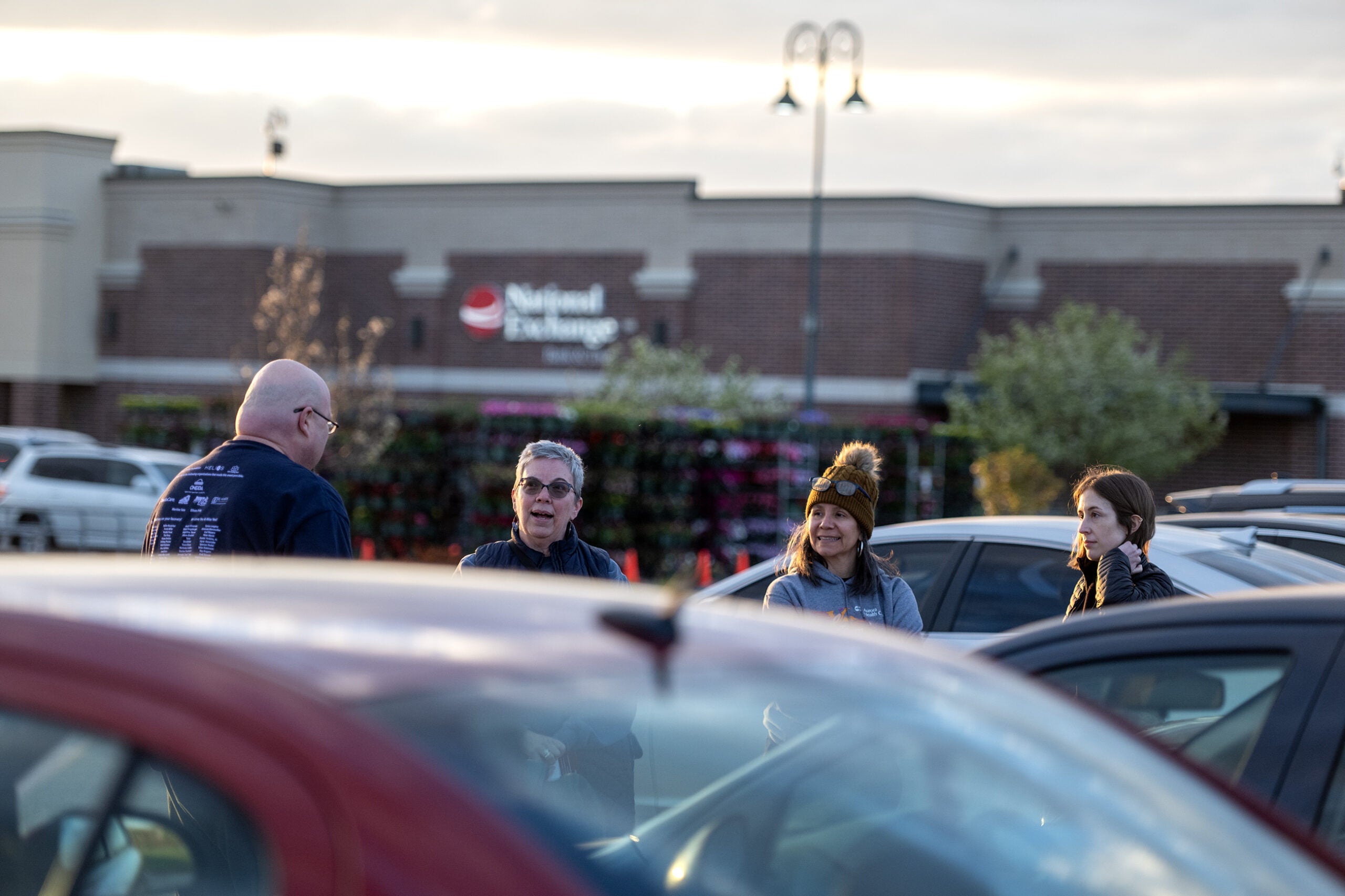 Four people stand talking in a parking lot near parked cars, with a National Exchange building in the background during daylight.