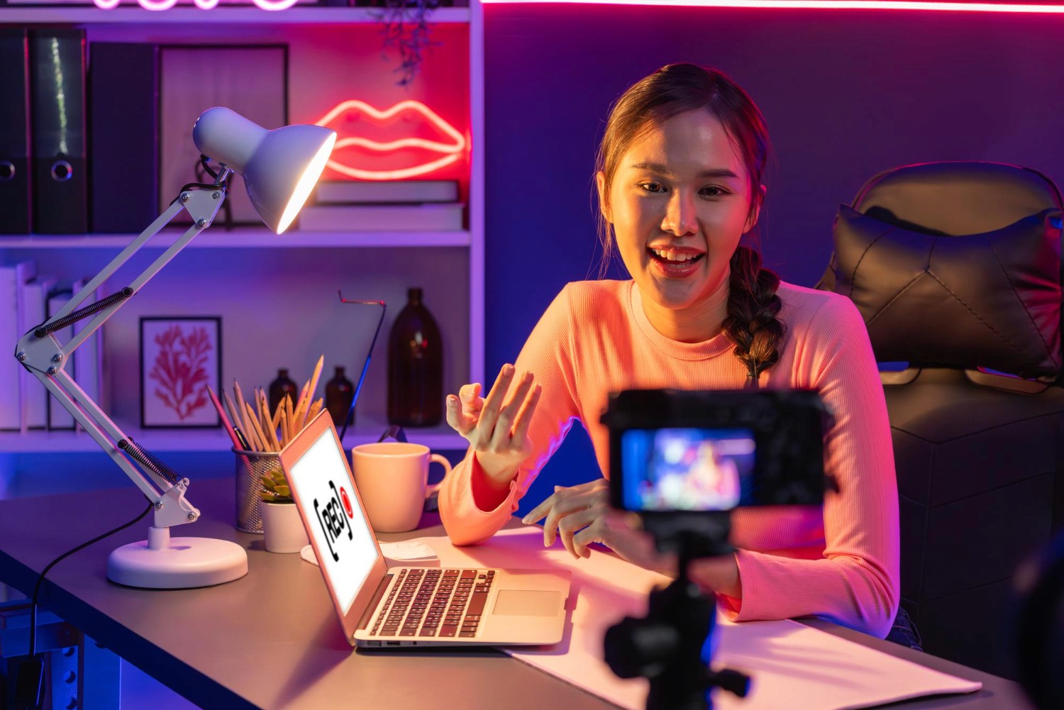 A woman at a desk using a laptop, accompanied by a bright neon sign that adds a modern touch to her workspace.