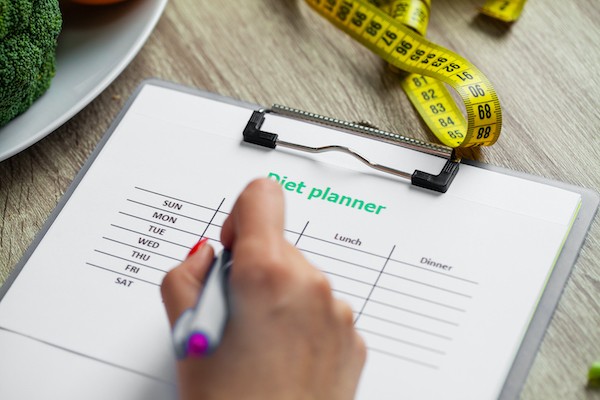 A hand holding a purple pen writes on a clipboard next to a plate of vegetables and measuring tape.