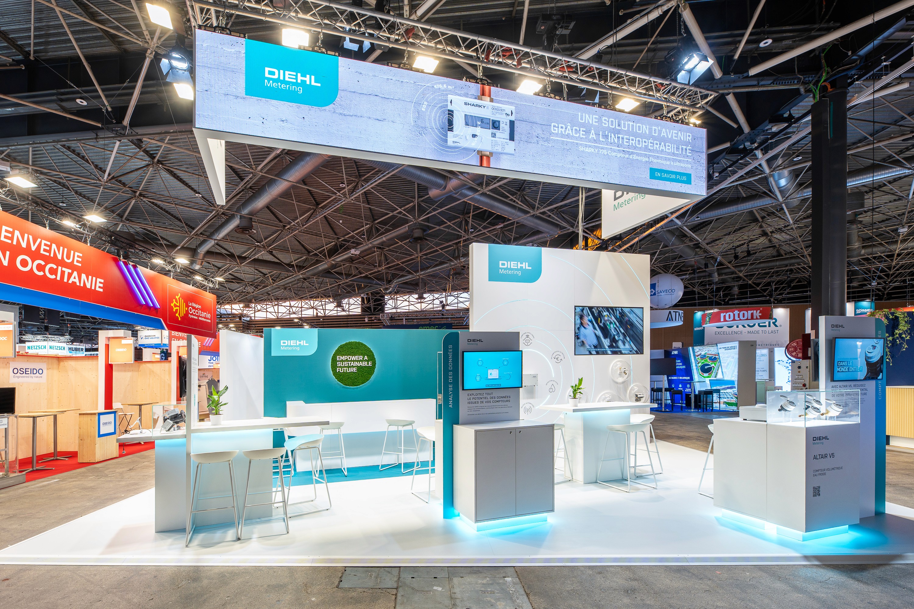 Stand Diehl Metering Pollutec 2025 conçu par Expace – vue d'ensemble du stand bipartite avec zone turquoise Empower a Sustainable Future, espace lounge et caissons de signalétique suspendus