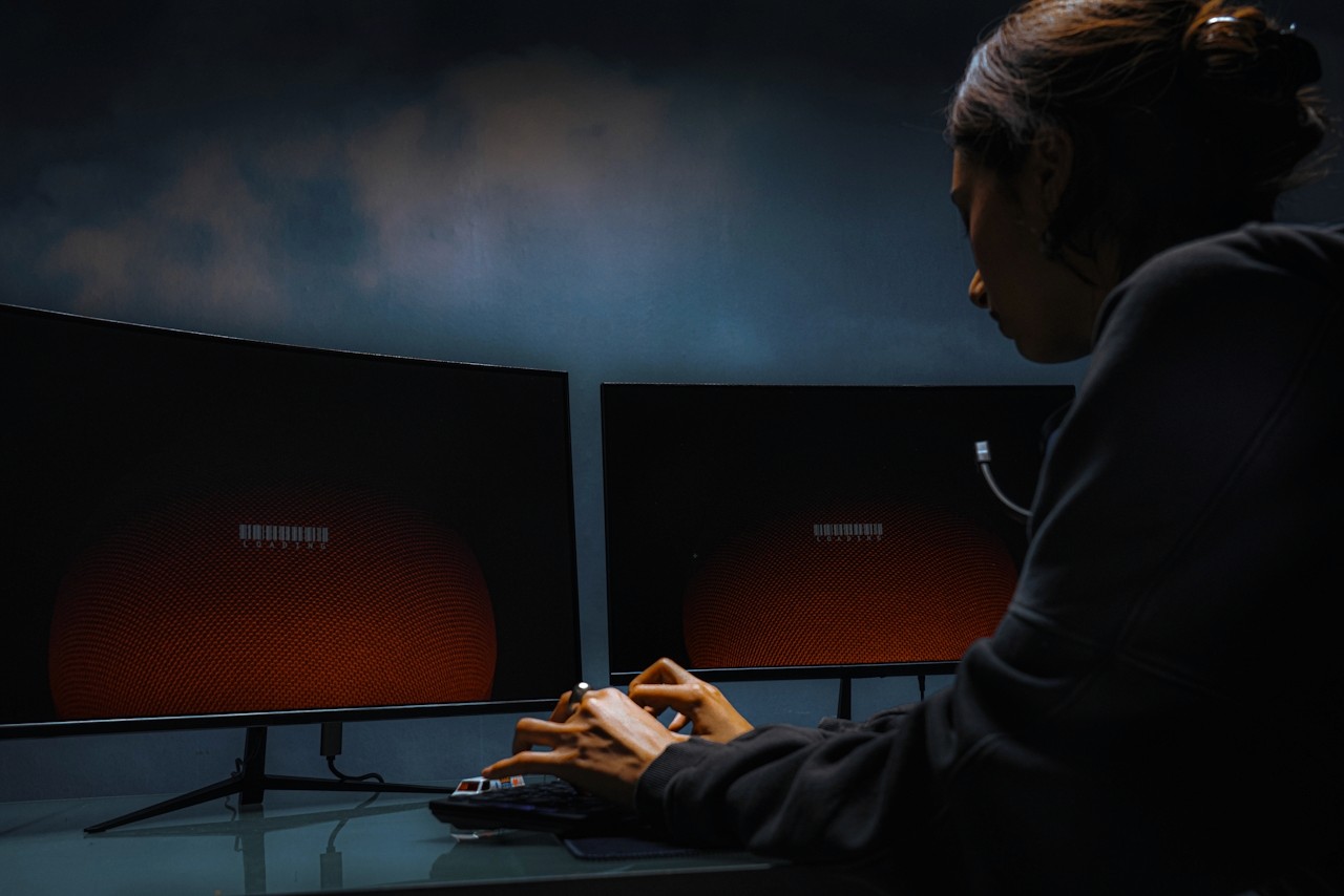 A person in a dark hoodie works at a keyboard in front of two large curved monitors glowing orange in a dim room.