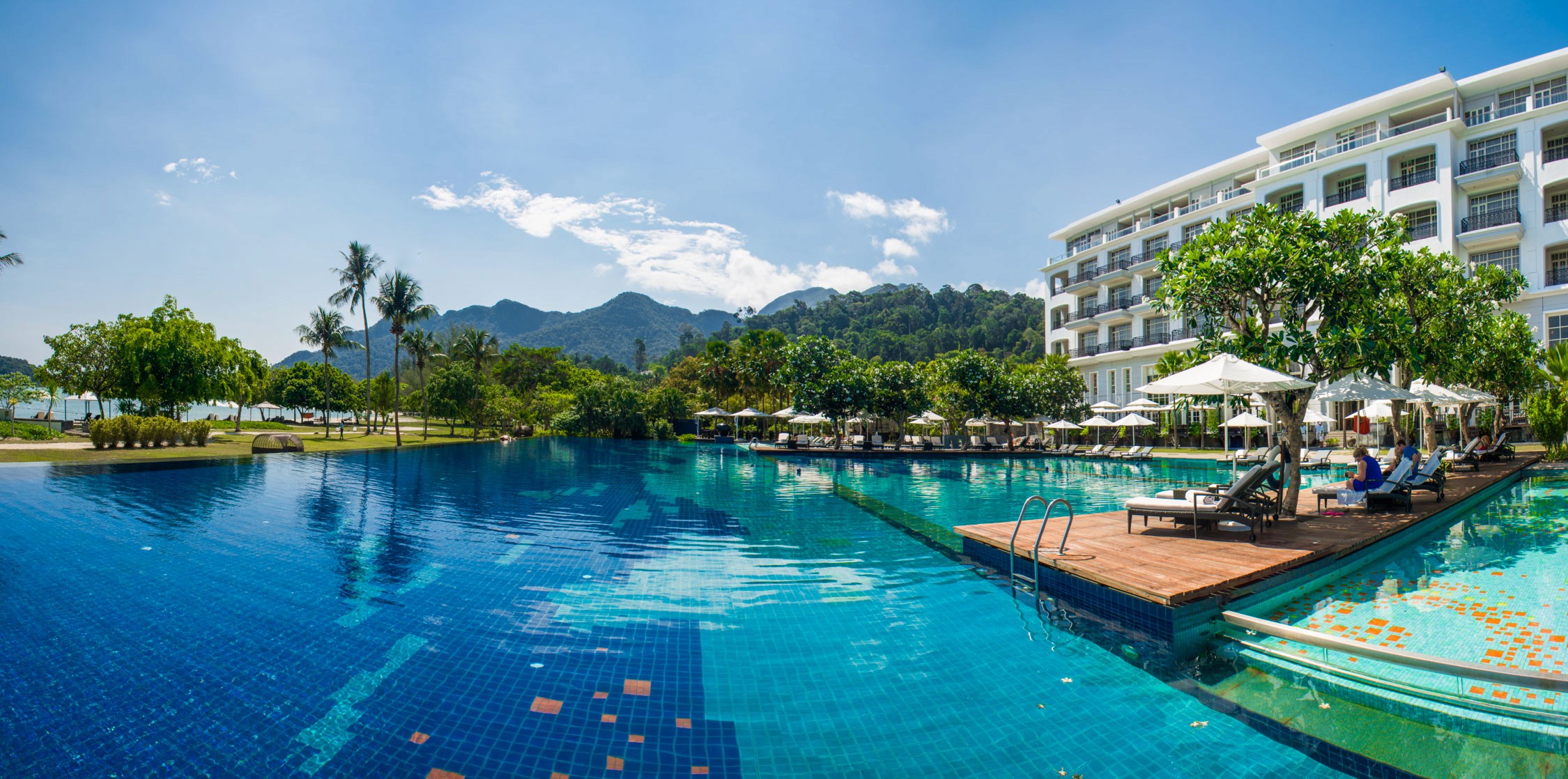 A resort swimming pool with blue tiles and lounge chairs overlooks a tropical landscape with mountains in the distance.