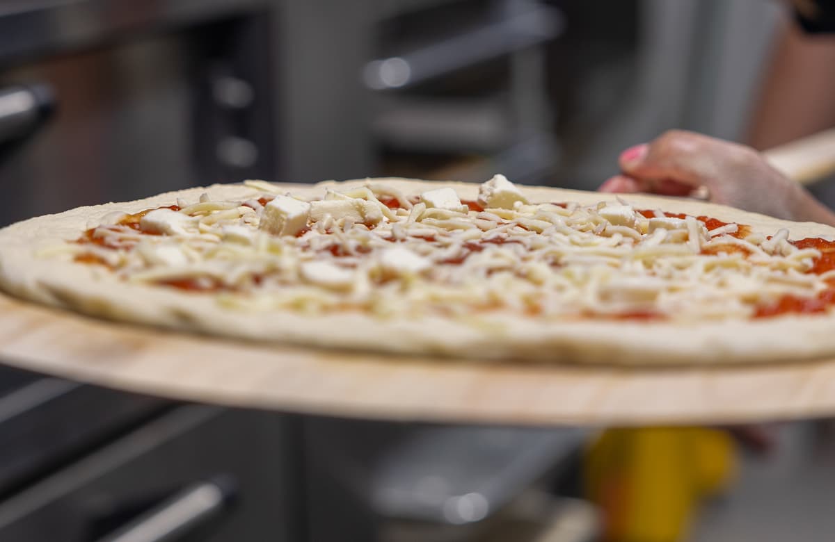 Unbaked pizza on a wooden peel topped with tomato sauce, shredded cheese, and cheese cubes, held by someone preparing to place it in the oven.