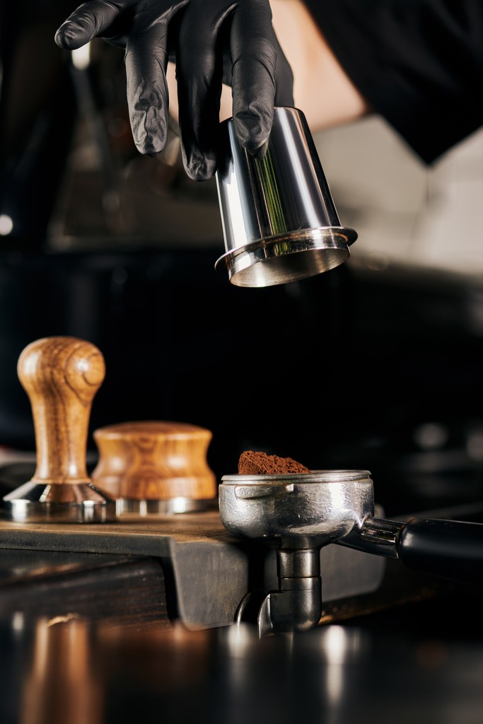 Barista using a metal dosing cup above a portafilter at an espresso bar, in a dark, moody setting.