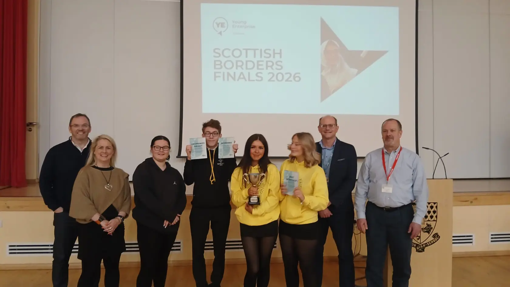 A group of eight people stands on a stage, smiling. Two hold a trophy and plaques. Behind them, a screen displays "Scottish Borders Finals 2026."