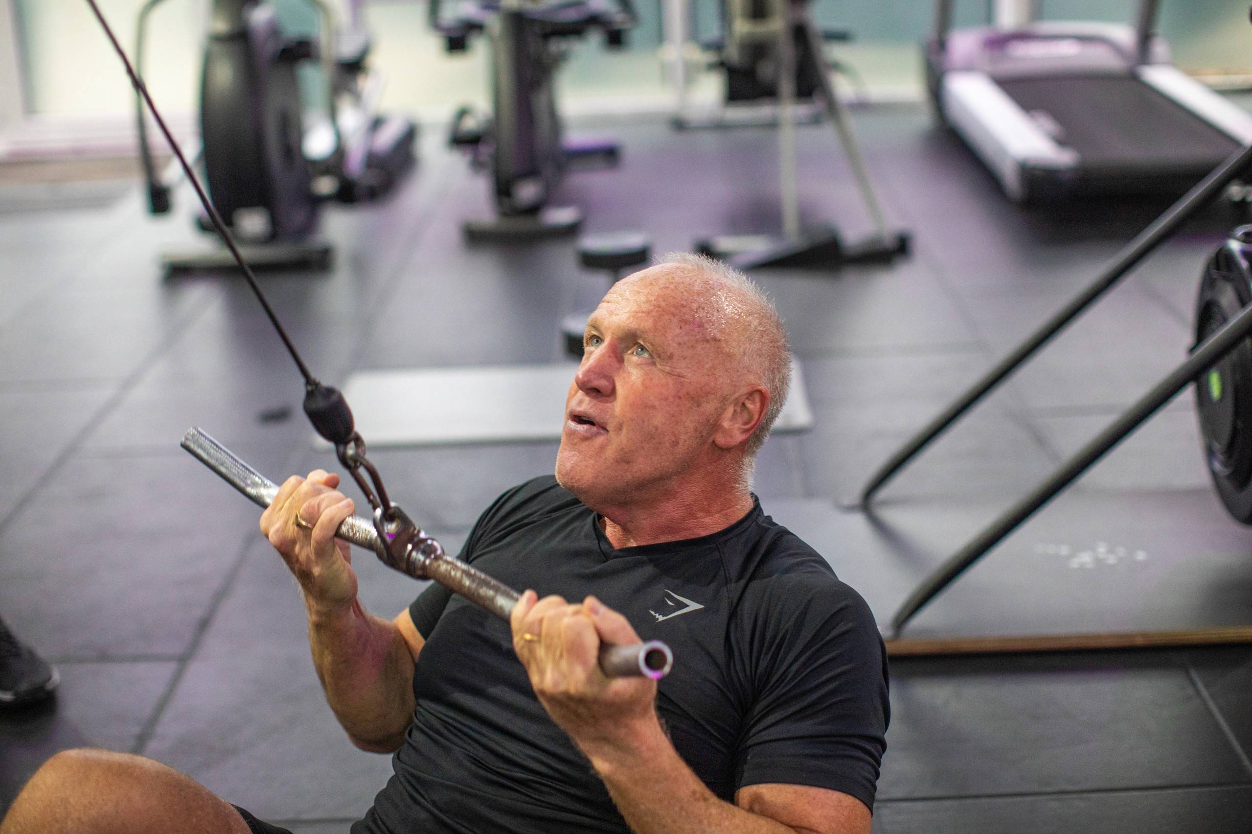 Man in a gym doing cable pull exercises, exerting effort. He's wearing a black shirt, surrounded by fitness equipment, conveying determination.