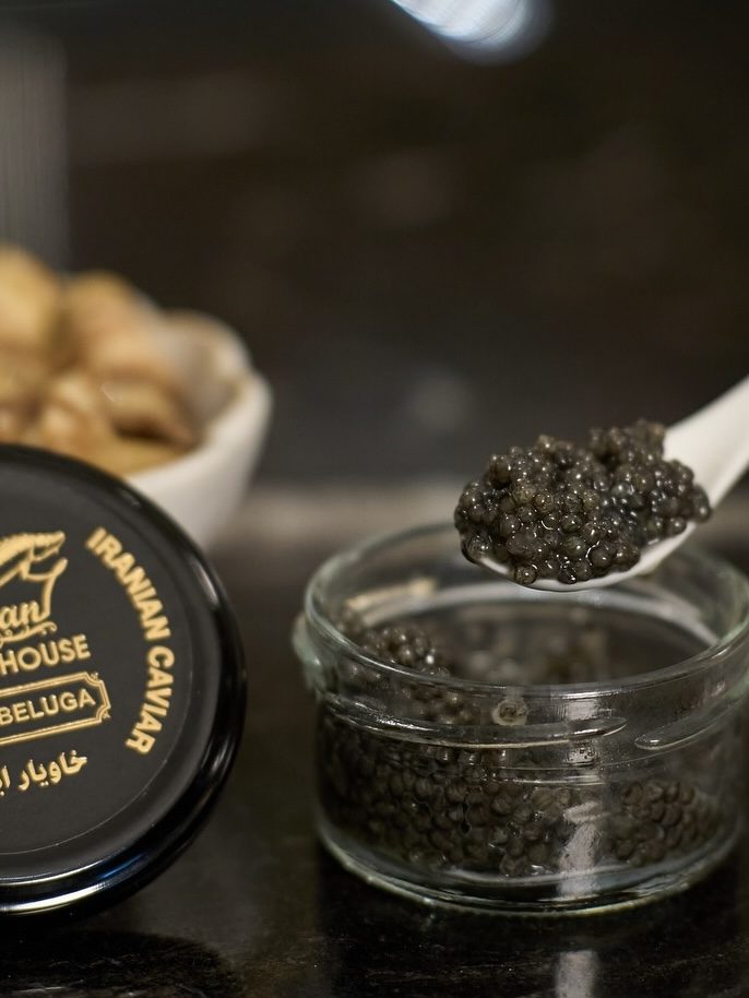 A hand holding a spoonful of caviar from a glass jar, beside a black tin labeled 'Caspian Caviar House - Royal Beluga', accompanied by a bowl of snacks and glasses of beverage on a dark surface.