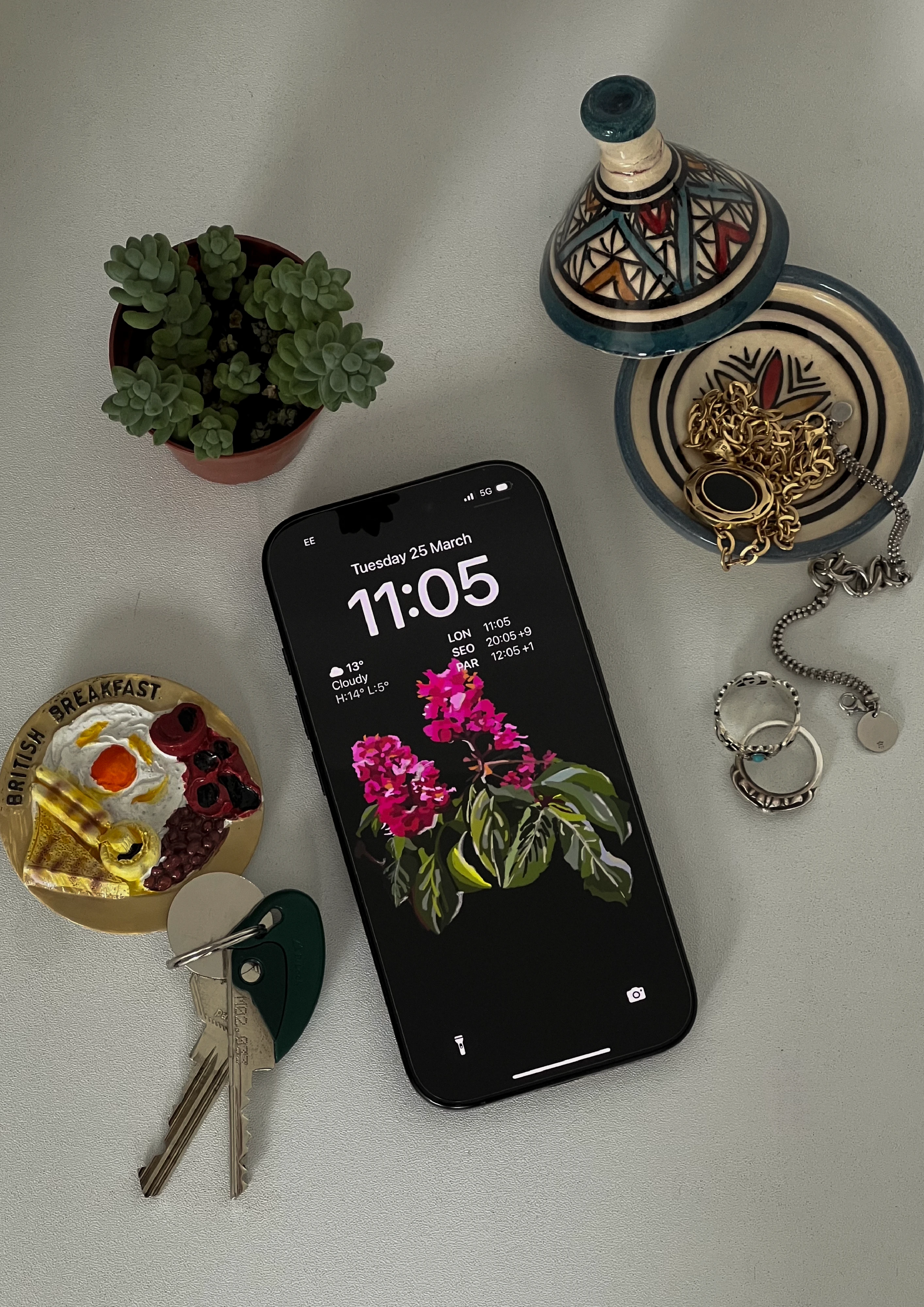 Phone on a pale surface showing a painted pink flowering branch as its lock screen, beside keys, rings, a small plant and a patterned lidded dish.