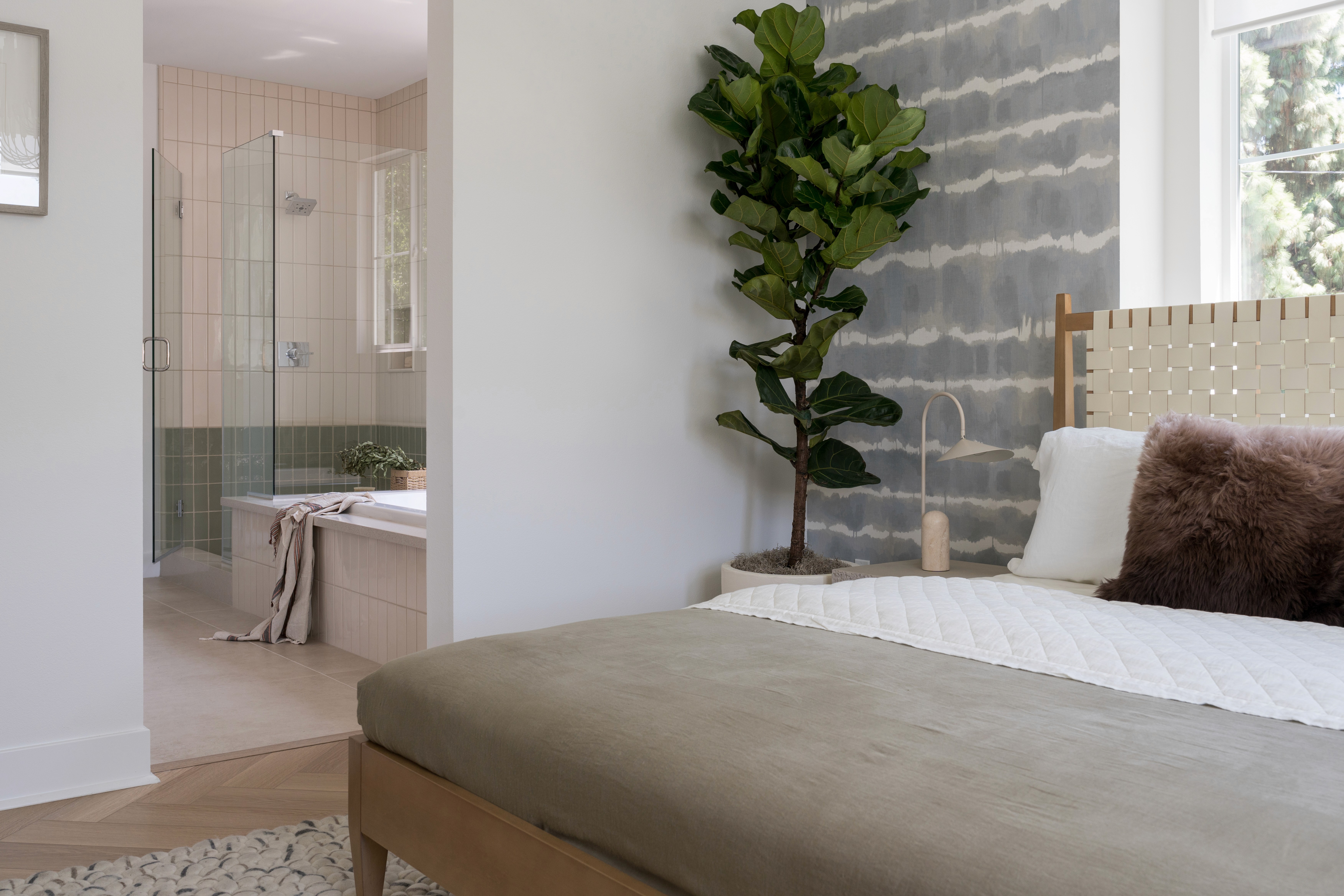 Playful bedroom and bathroom with wood bed frame, woven rug, chevron wood floors with off-white tile in bathroom, grey and white accent wall by Jubilee Interiors in Arcadia, California