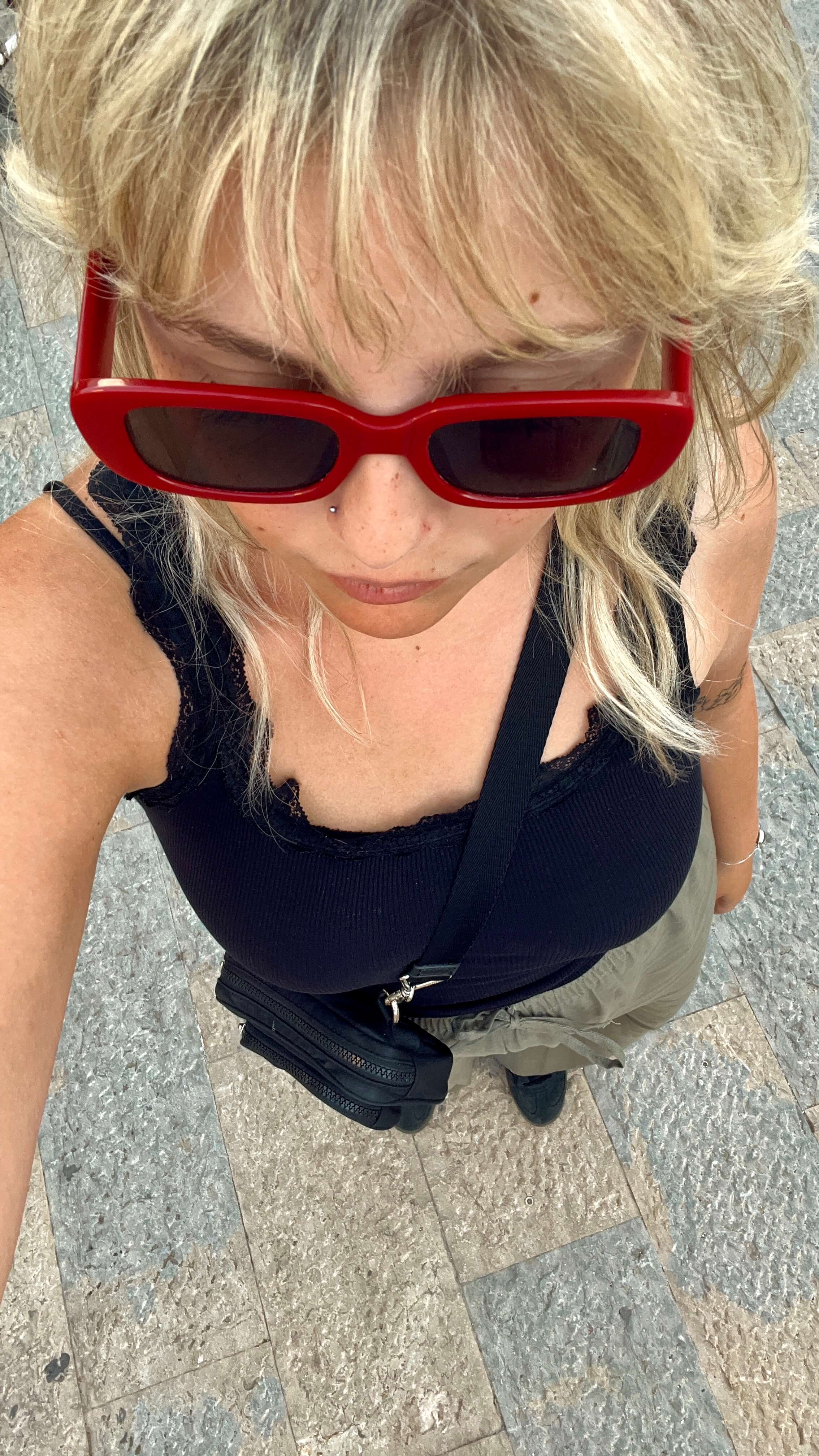 Young Blonde woman with bangs wearing red sunglasses taking a selfie from high up so that you can see her whole body