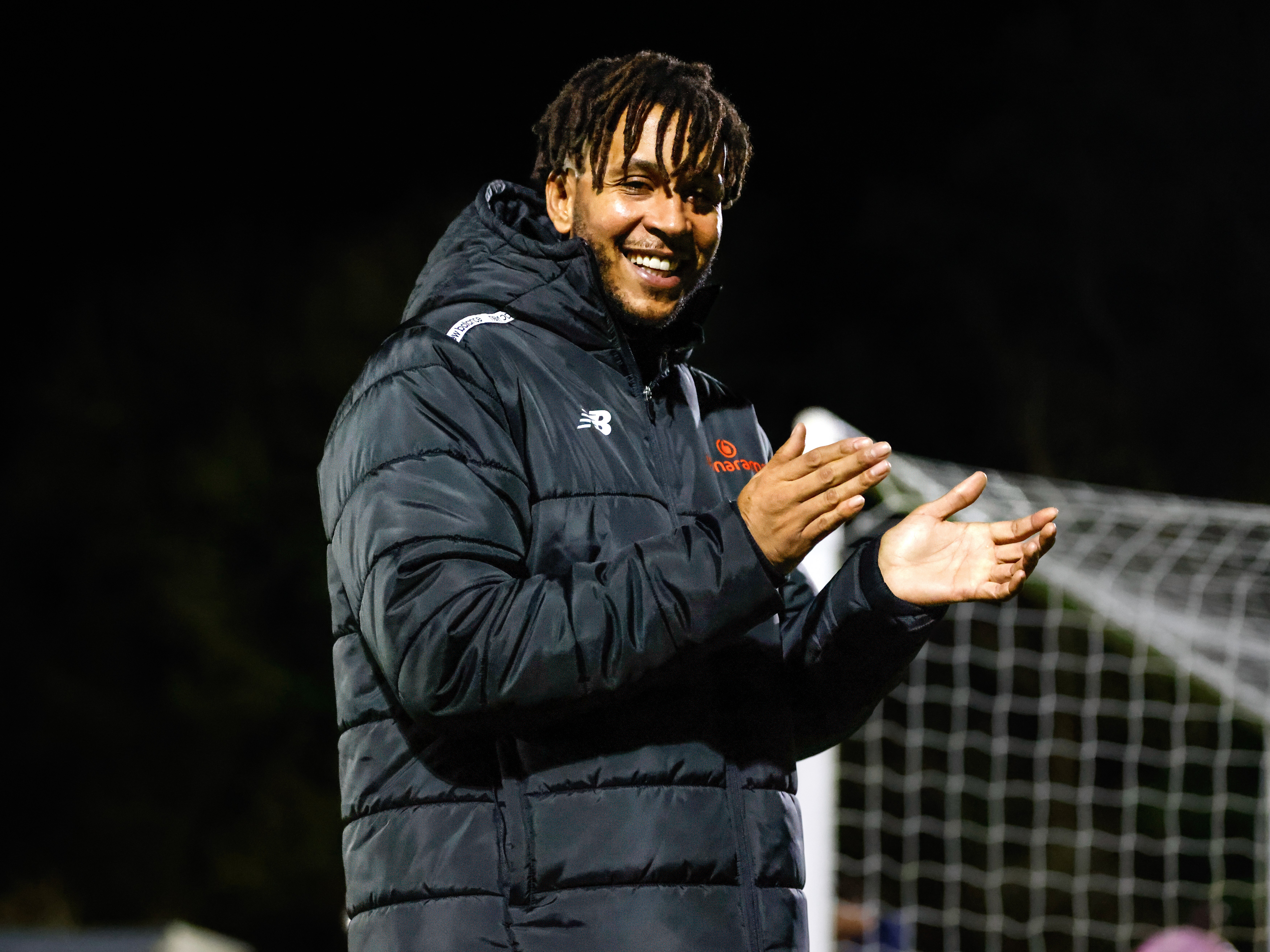 A person wearing a dark padded jacket stands by a soccer goal, smiling and clapping hands together during a nighttime outdoor sports event.