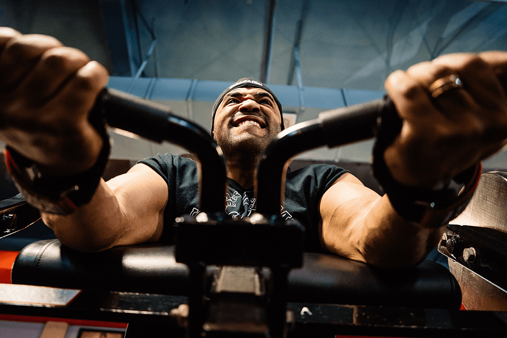 A man with a broad smile is seated on a black weight machine, gripping the handles with both hands, wearing a dark green t-shirt and a dark baseball cap. The background is blurred, showcasing a blue-gray wall.