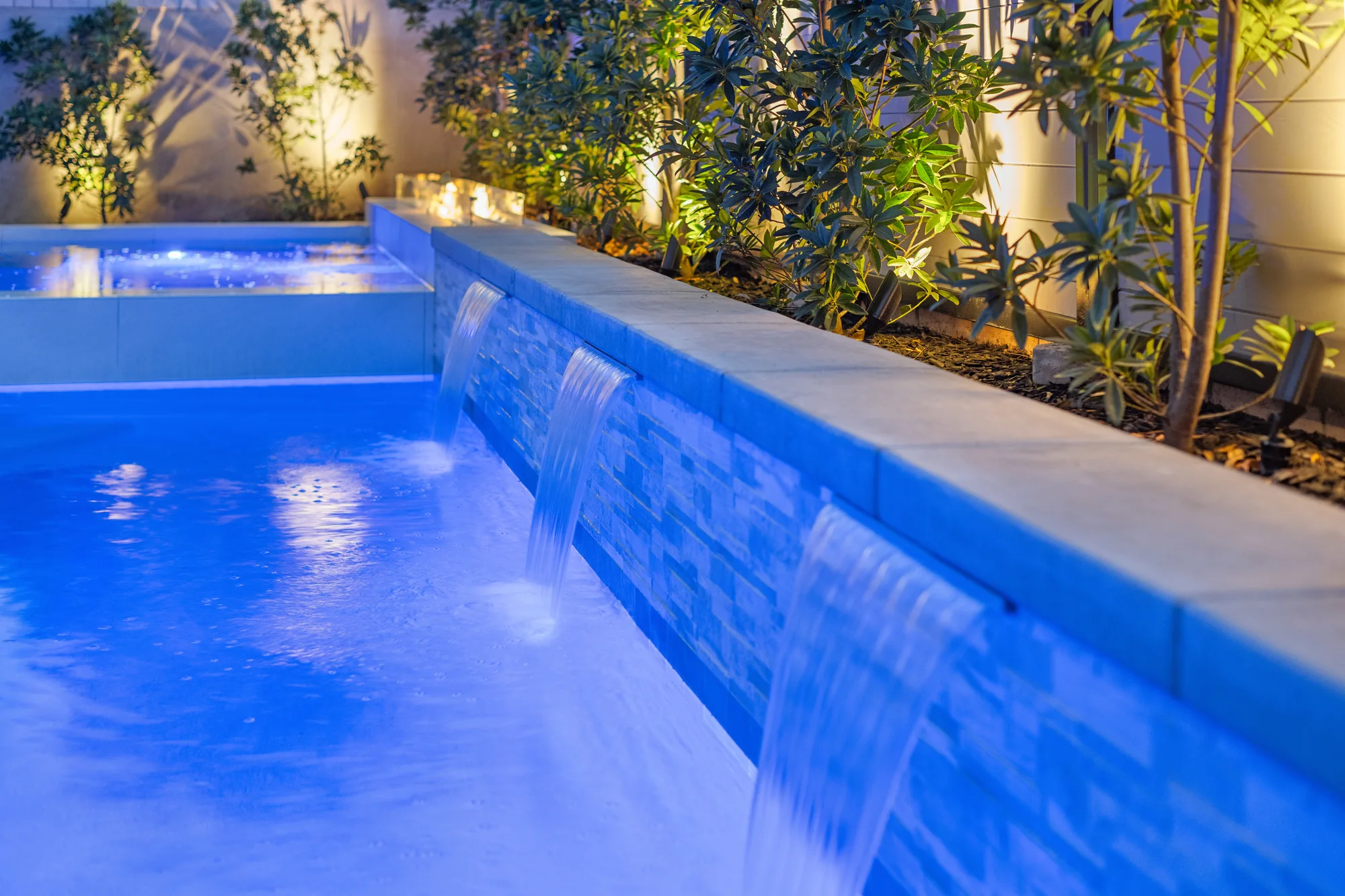 Water flowing from a stone-faced wall into a blue-lit swimming pool.