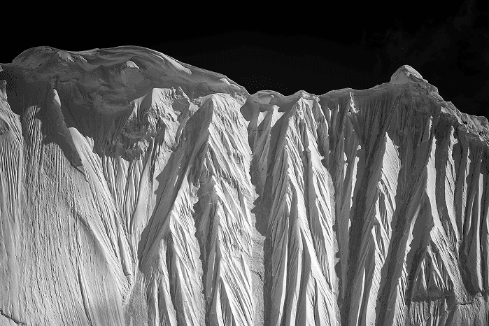 Czarno-biała fotografia ośnieżonych gór z wyraźnymi żłobieniami i dramatycznym światłocieniem na tle ciemnego nieba