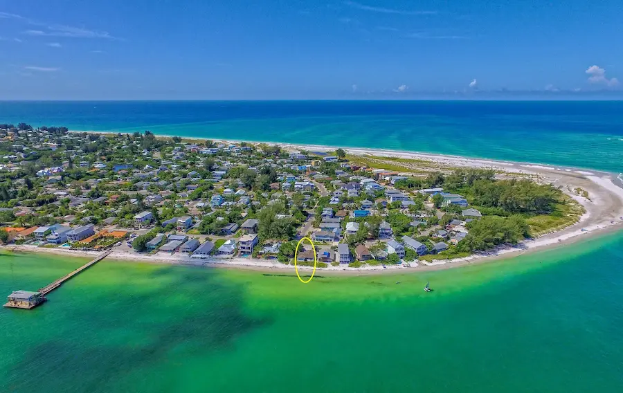 Aerial view of Anna Maria Island, FL, showcasing a vibrant tropical coastline with turquoise waters, white sandy beaches, and a community of colorful beachfront homes under a clear blue sky.