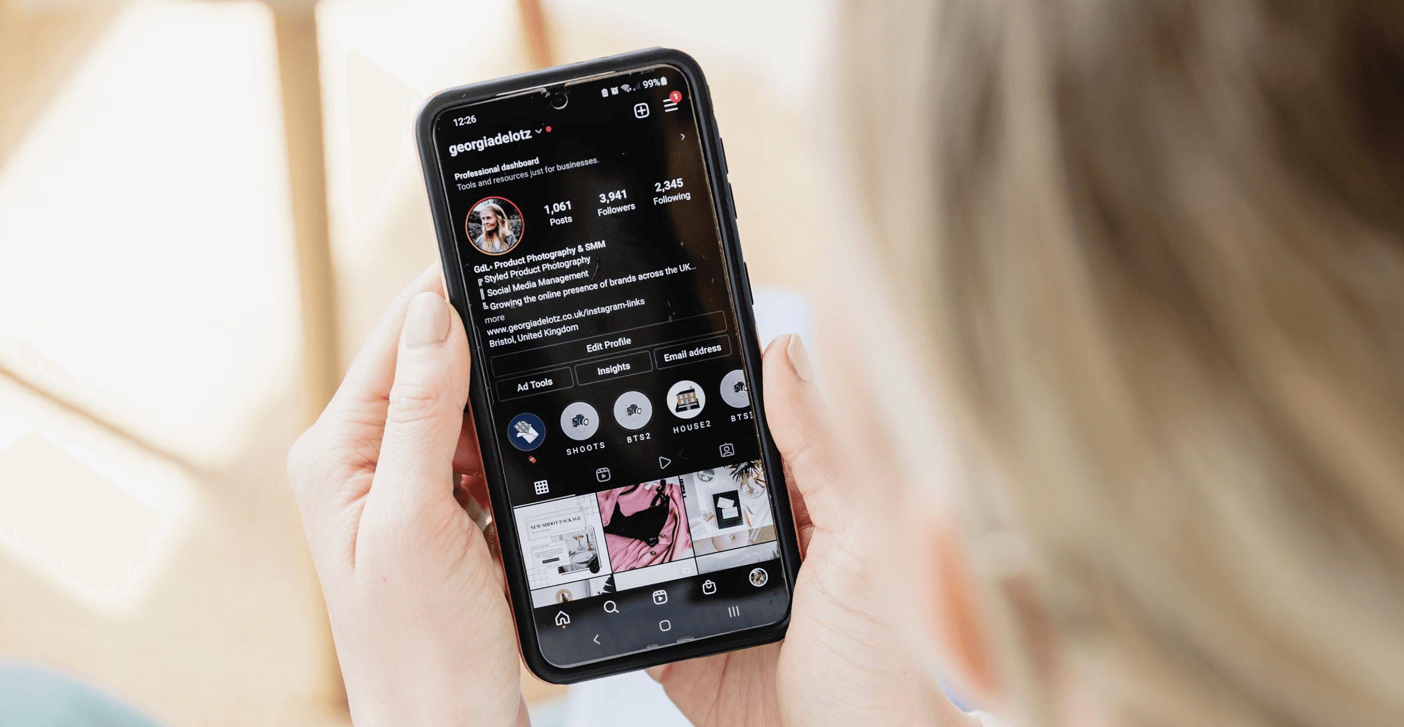 A person holds a smartphone displaying a social media profile page, with a table holding cups in the background.