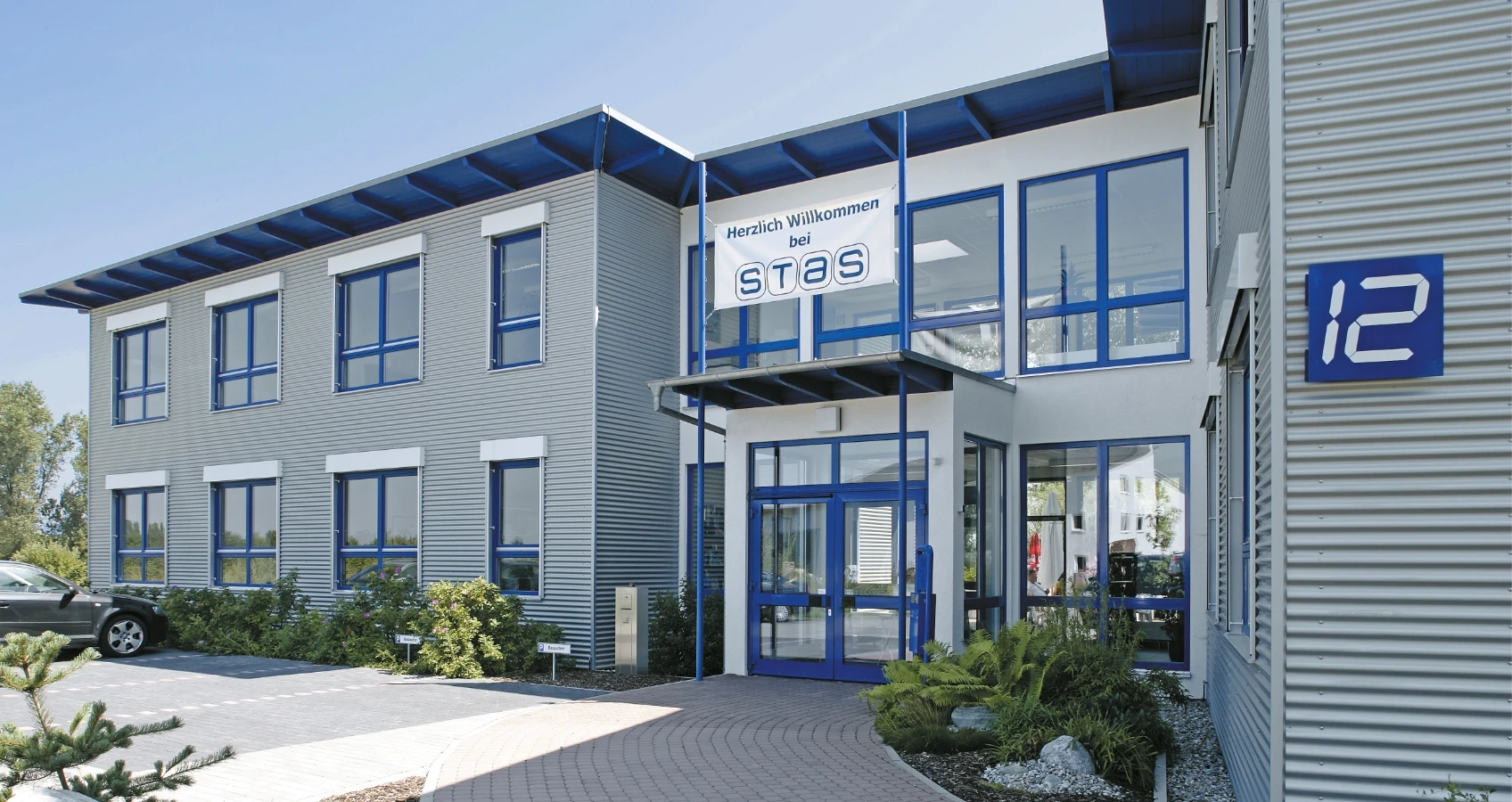 Modernes Firmengebäude mit grauer Wellblechfassade, blauen Fenstern, einem blauen Vordach und einem Willkommensbanner für STAS.