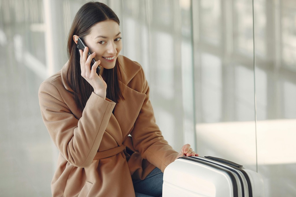 A smiling newcomer arriving in Canada sitting with her luggage at the airport, happily making a call on her smartphone; representing a traveler who successfully avoided expensive Canadian cell phone provider kiosks by using an affordable travel eSIM.