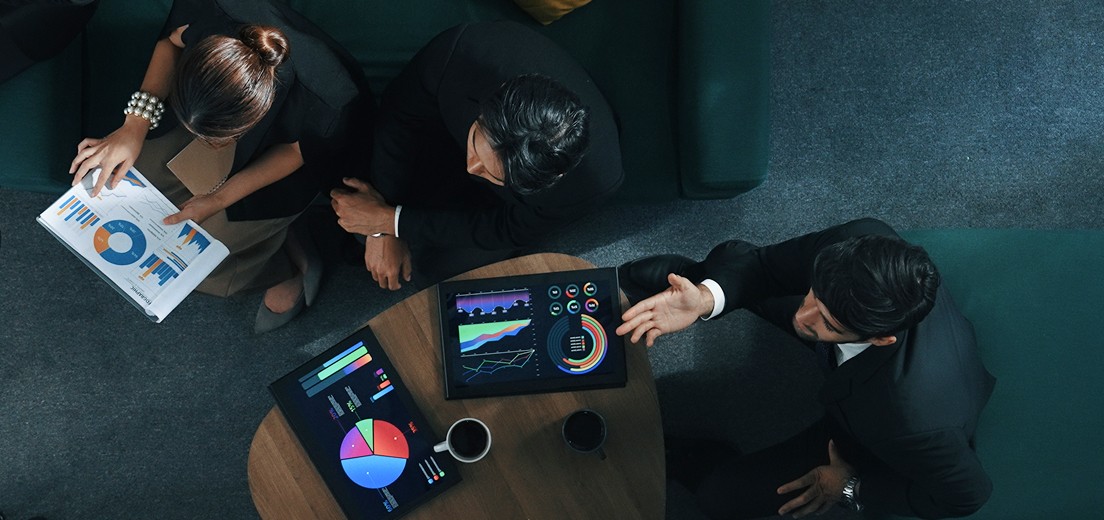 A high-angle view of a business meeting shows three people sitting around a wooden table with several digital tablets and papers displaying colorful charts and graphs, indicating a focus on data analysis and strategy discussion.