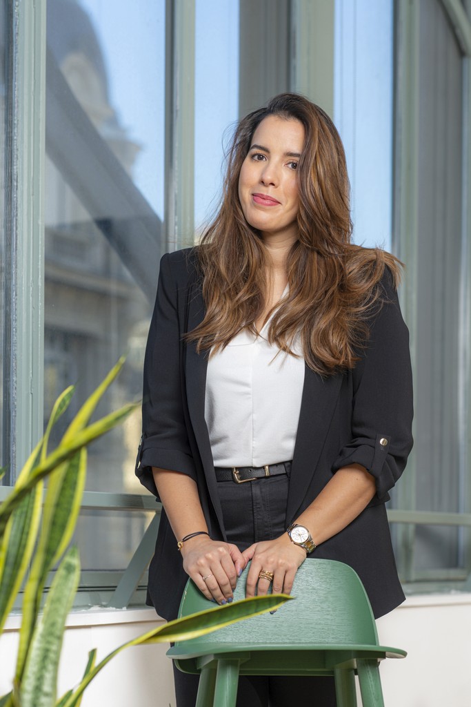 Young person with curly hair wearing a rust-colored blazer and black turtleneck, looking confidently at the camera against a neutral background.