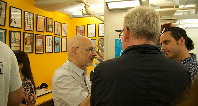A group of people talking to each other while standing in an exhibition room with some paintings on the wall.