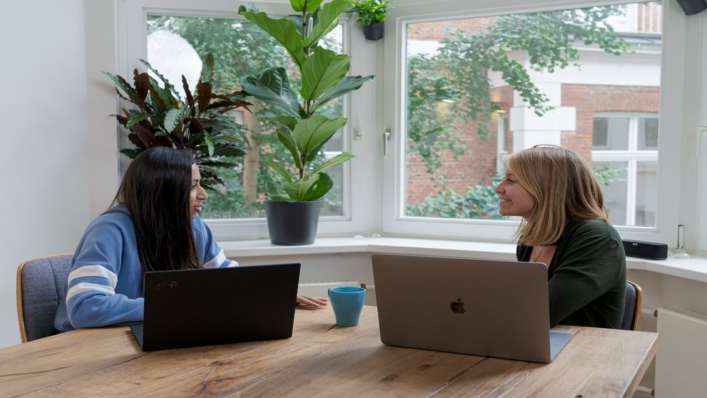 Een vrouw en iemand zitten aan een tafel met laptops, omringd door groen in een lichte, moderne werkruimte.