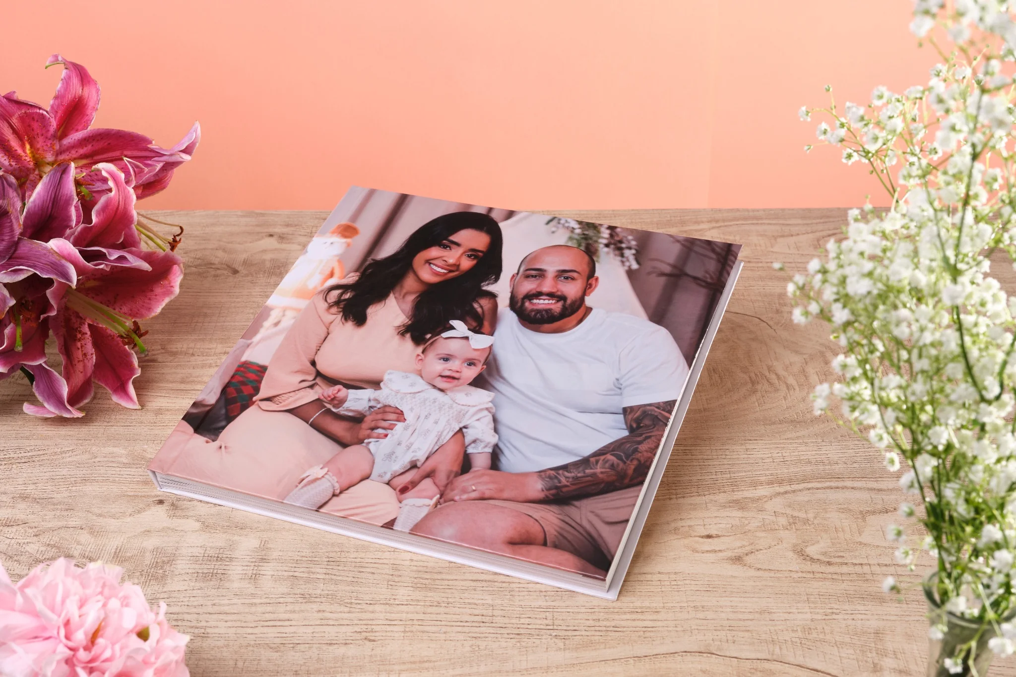 Mãos segurando fotolivro com capa personalizada das Maldivas, com casal ao pôr do sol e flores decorativas ao redor, sobre mesa de madeira clara