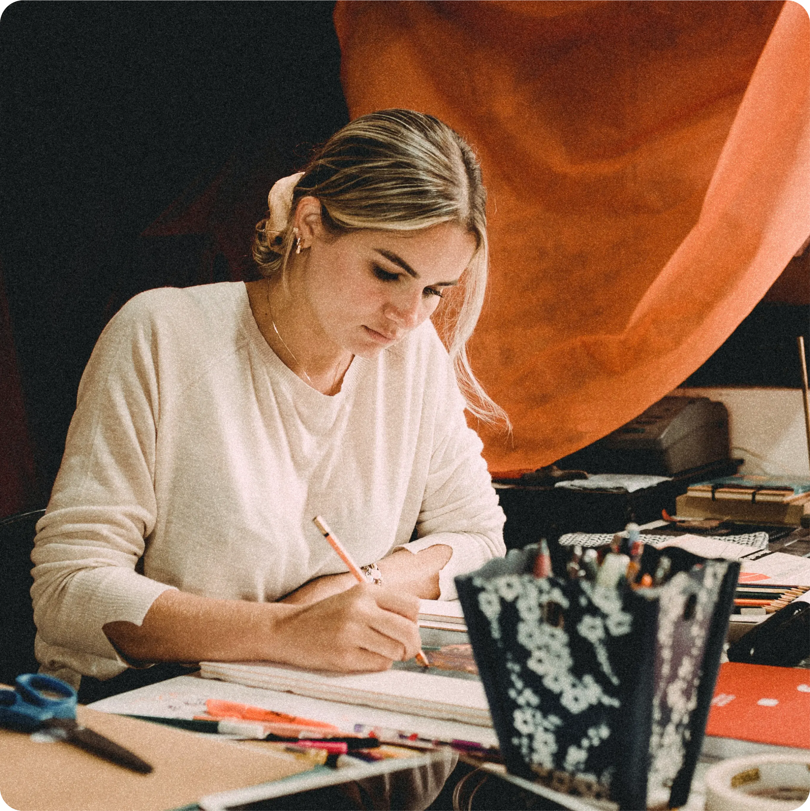 A woman sits at a cluttered desk, focusing intently on her work. She is writing in a notebook with a pencil, surrounded by various art supplies, including markers, scissors, and a container holding more pencils. She wears a light-colored sweater and has her blonde hair tied back with a hairband. The background features an orange fabric draped behind her.