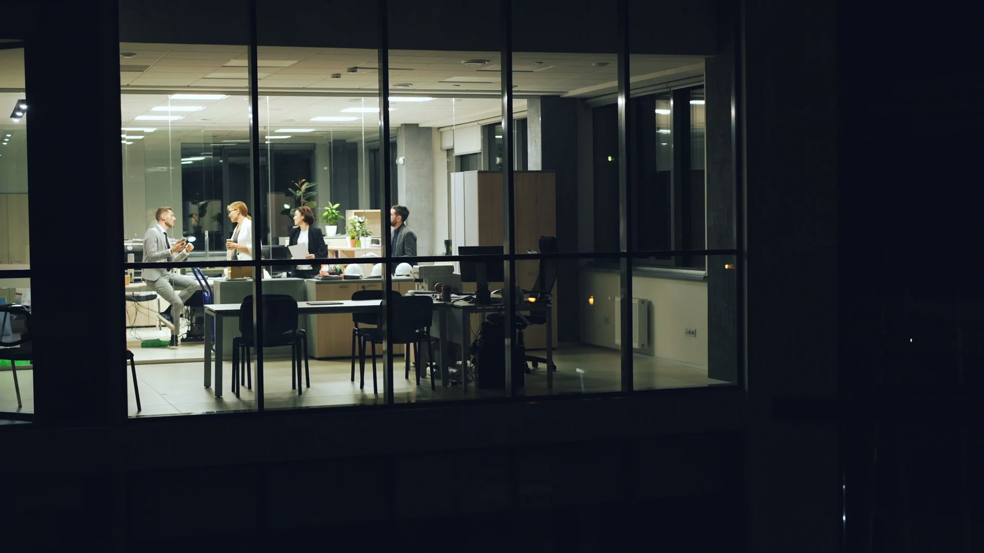 A wide, exterior night view through large glass windows of a brightly lit corporate office where a small team of professionals is working late.