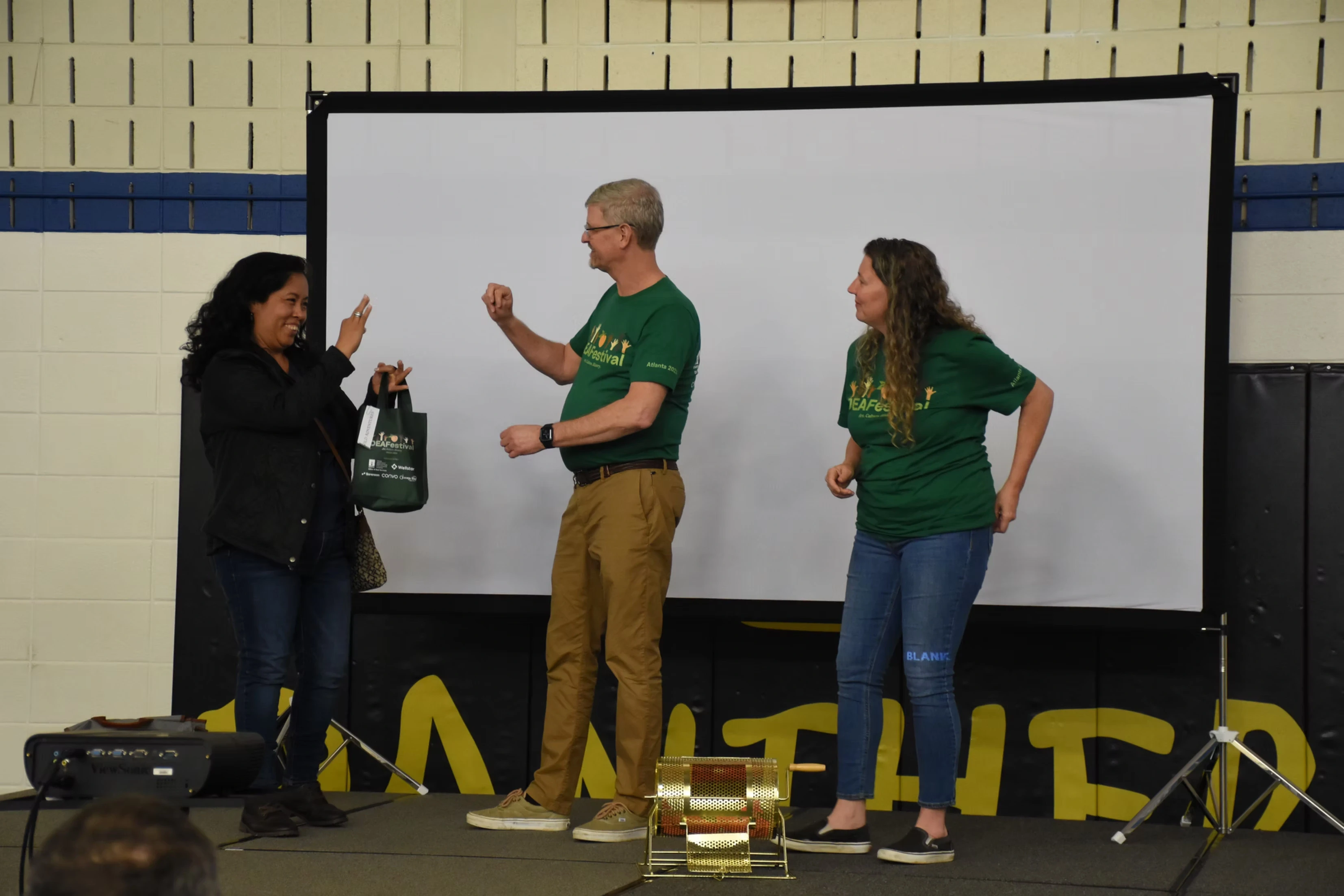 Three people on a stage, one handing a gift bag to another, during a presentation or event.