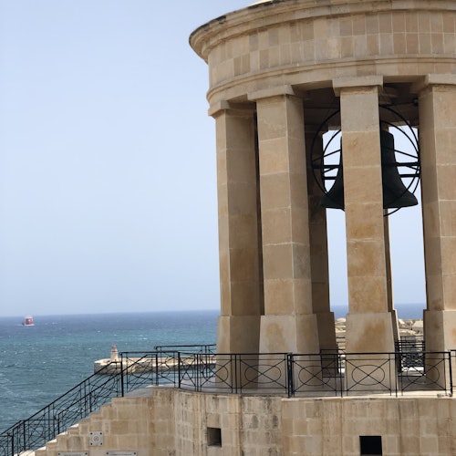 A stone structure with columns houses a large bell overlooking a blue sea and distant horizon. A red buoy is visible in the water.