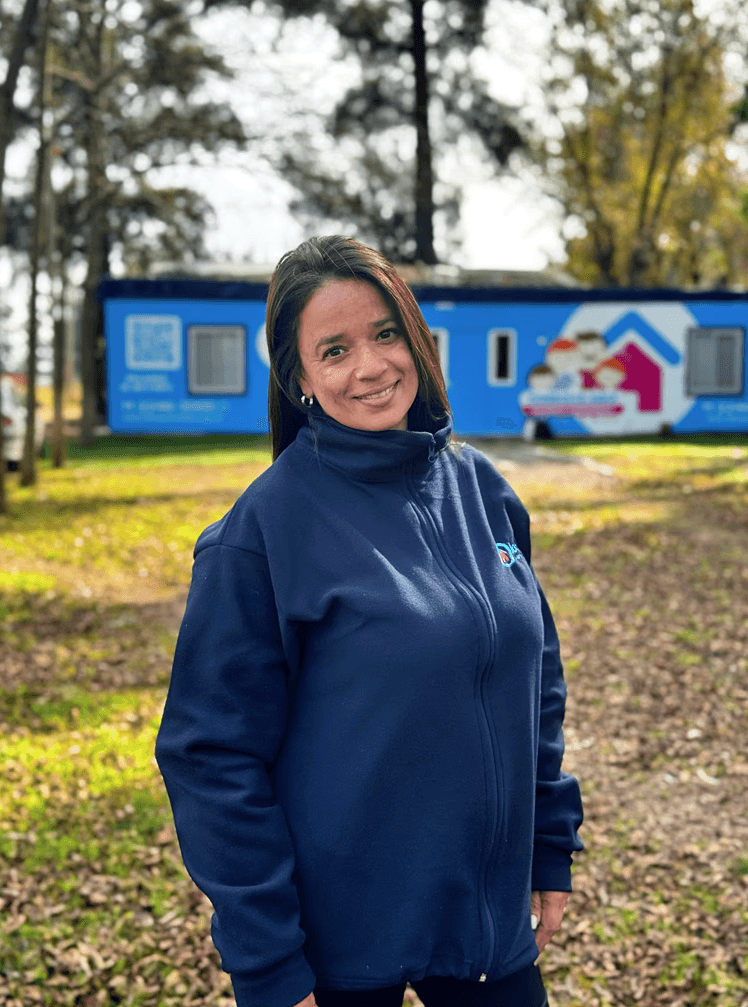 Smiling young woman with long hair standing against a dark green background, holding a finger to her chin.