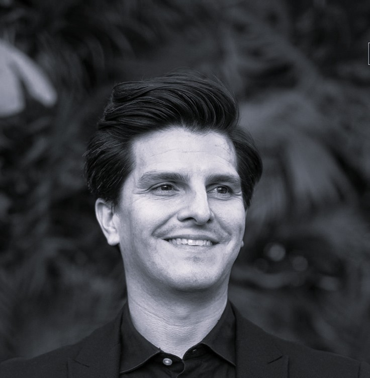 A young person with curly hair smiles at the camera while sitting in front of a laptop against a dark background.