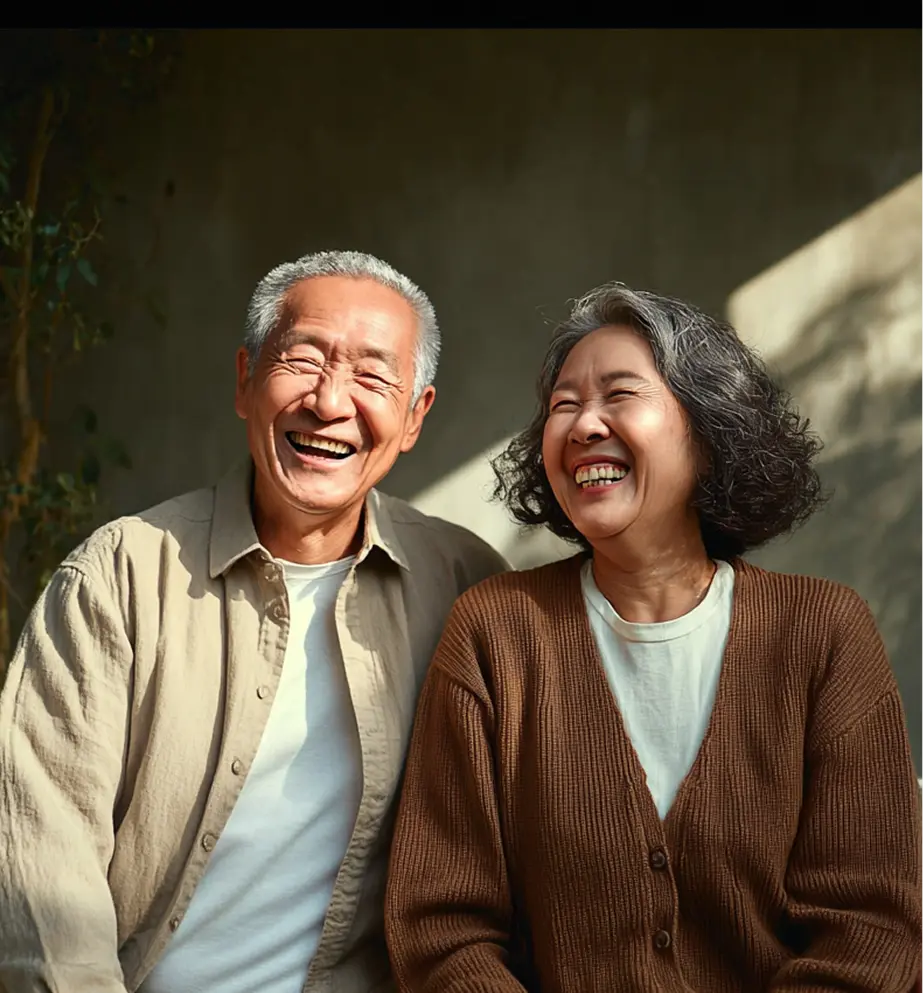 Older couple sitting together outdoors, laughing warmly in soft sunlight, wearing casual neutral-toned clothing with relaxed, happy expressions.