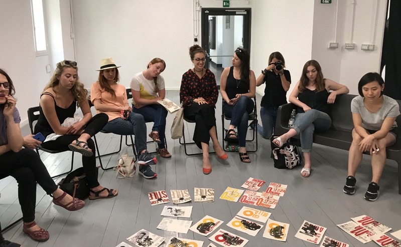 A photo of a group of people sitting on chairs in a circle manner, while looking at some cover book artwork set on the floor in the middle.