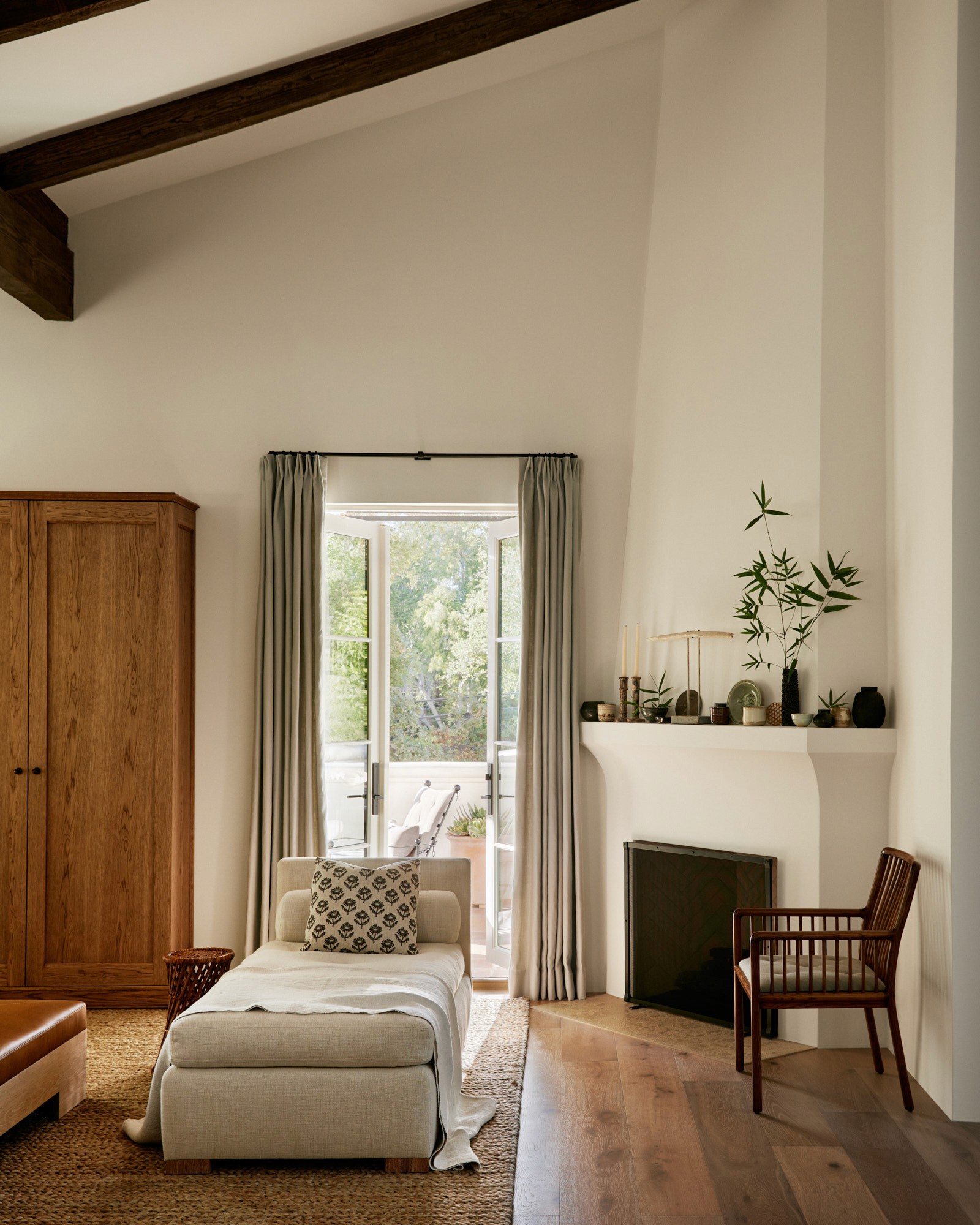 A living room corner with a white plaster fireplace, its mantel arranged with ceramics, candles, and a cutting of greenery. A chaise upholstered in natural linen and a spindle-back wood chair flank the hearth. French doors open to an outdoor terrace beyond.