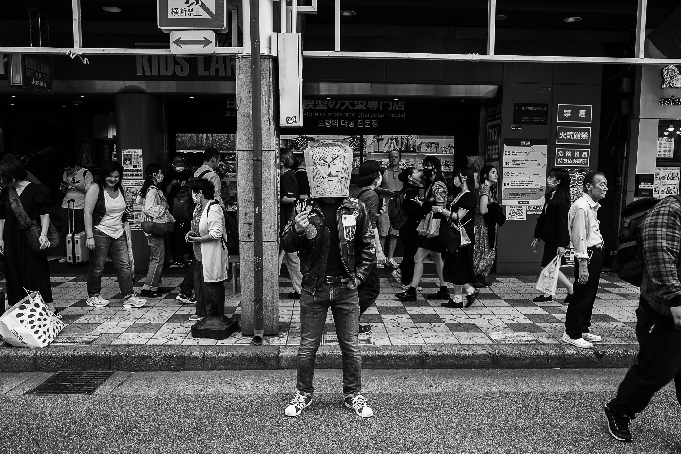 Black and white street photograph of a man standing in front of a shop in a busy city street.