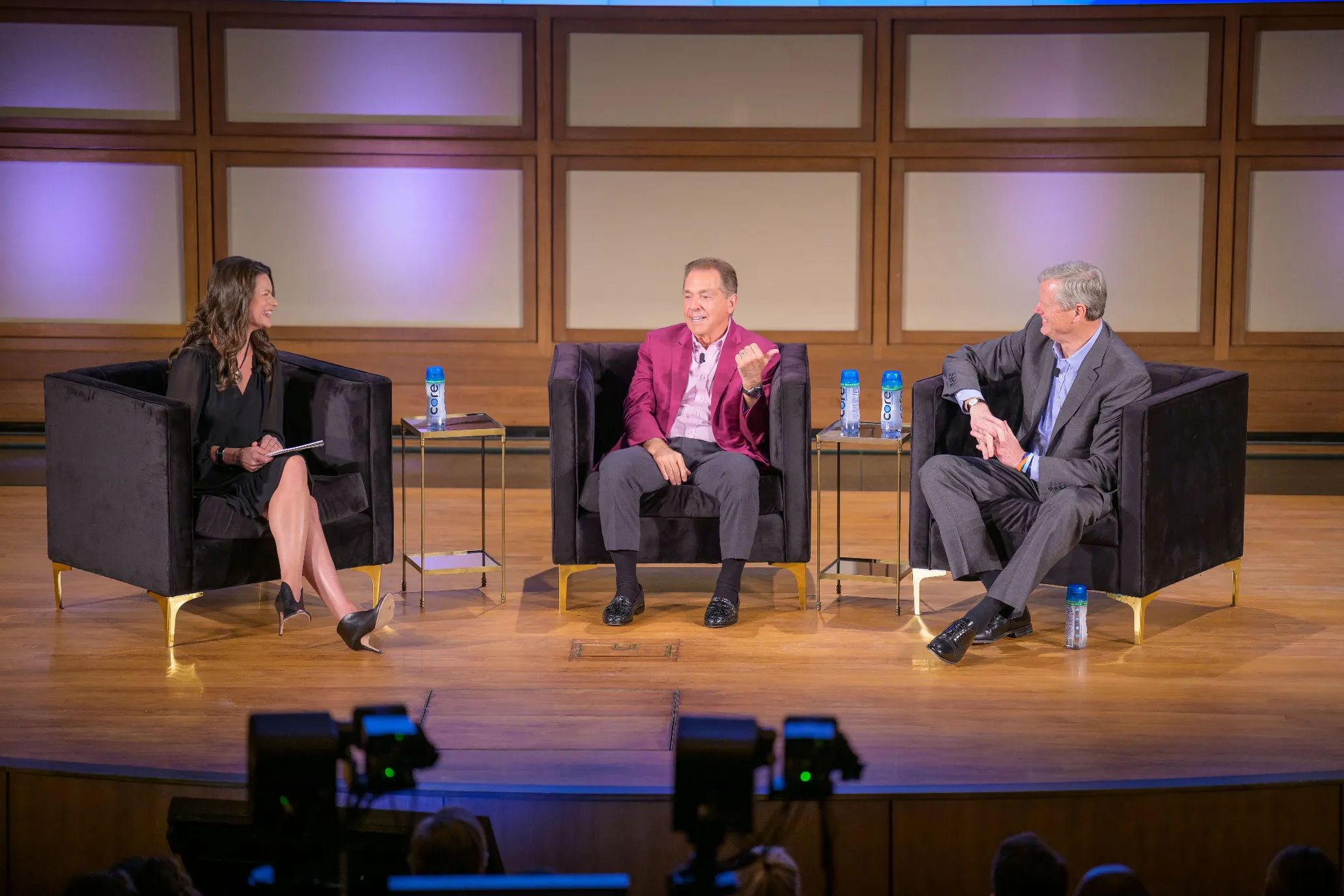Three people sit on stage in armchairs, engaged in a panel discussion. Heather Dinich on the left holds papers, while the two men on the right, one in a maroon jacket, are talking and smiling. Bottled water sits on side tables beside them.