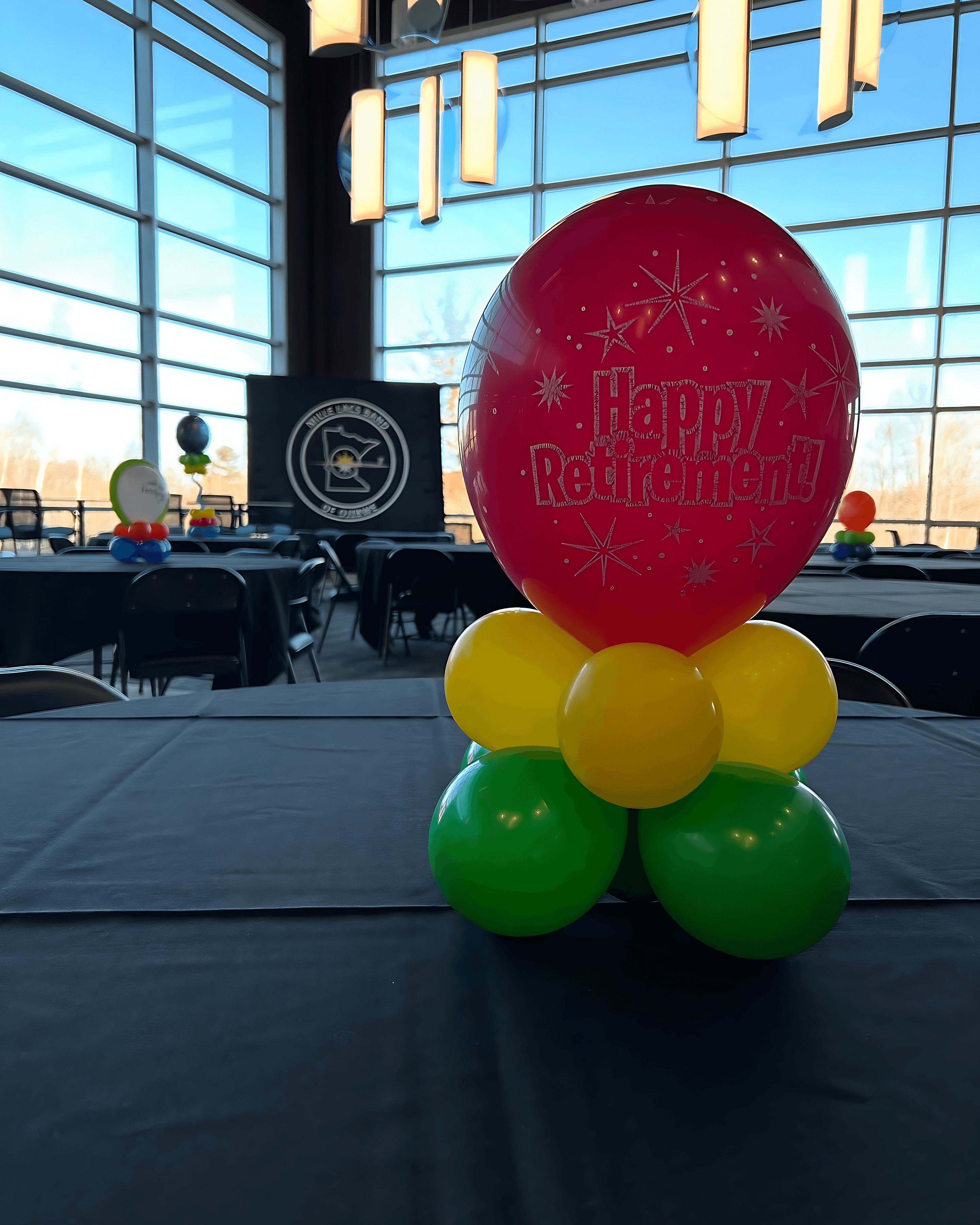 Red and white balloon centerpiece with green base reading Happy Birthday displayed at an indoor event by Be Bold Balloons