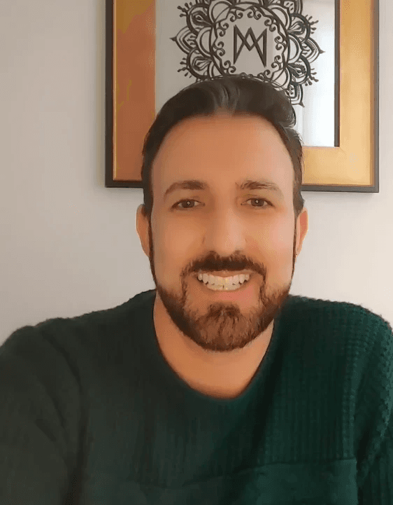 Smiling female therapist with short gray hair, wearing a brown top and a purple shawl, surrounded by a cozy background.