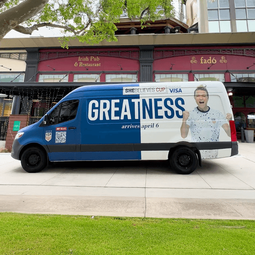 A blue and white SheBelieves Cup van with GREATNESS arrives april 6 written on the side, parked in front of an Irish pub and restaurant called Fadó. A cheering woman is also featured on the van.