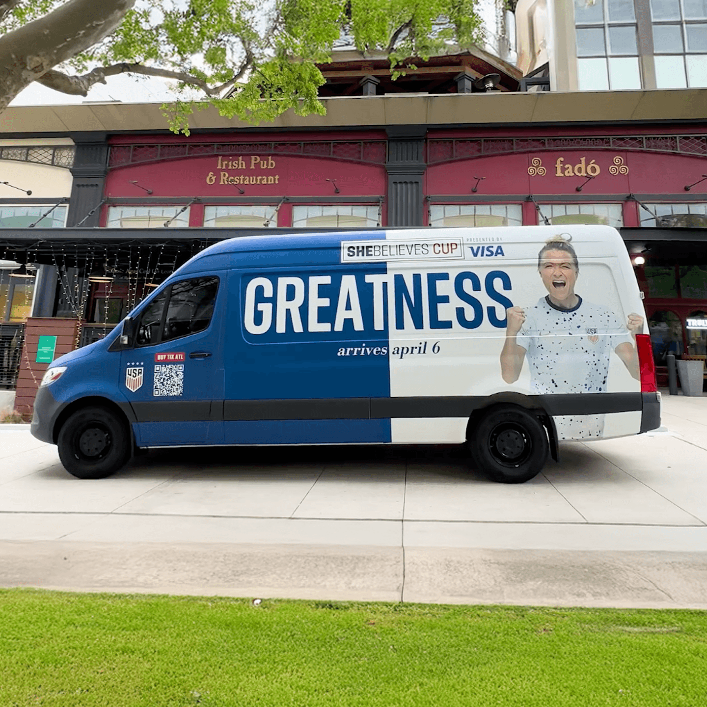 A blue and white SheBelieves Cup van with GREATNESS arrives april 6 written on the side, parked in front of an Irish pub and restaurant called Fadó. A cheering woman is also featured on the van.